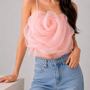 Pu&ograve; includere: Un top corto rosa chiaro con un grande design a forma di rosa realizzato in tessuto a strati. Il top ha sottili spalline ed &egrave; abbinato a jeans di denim blu chiaro.