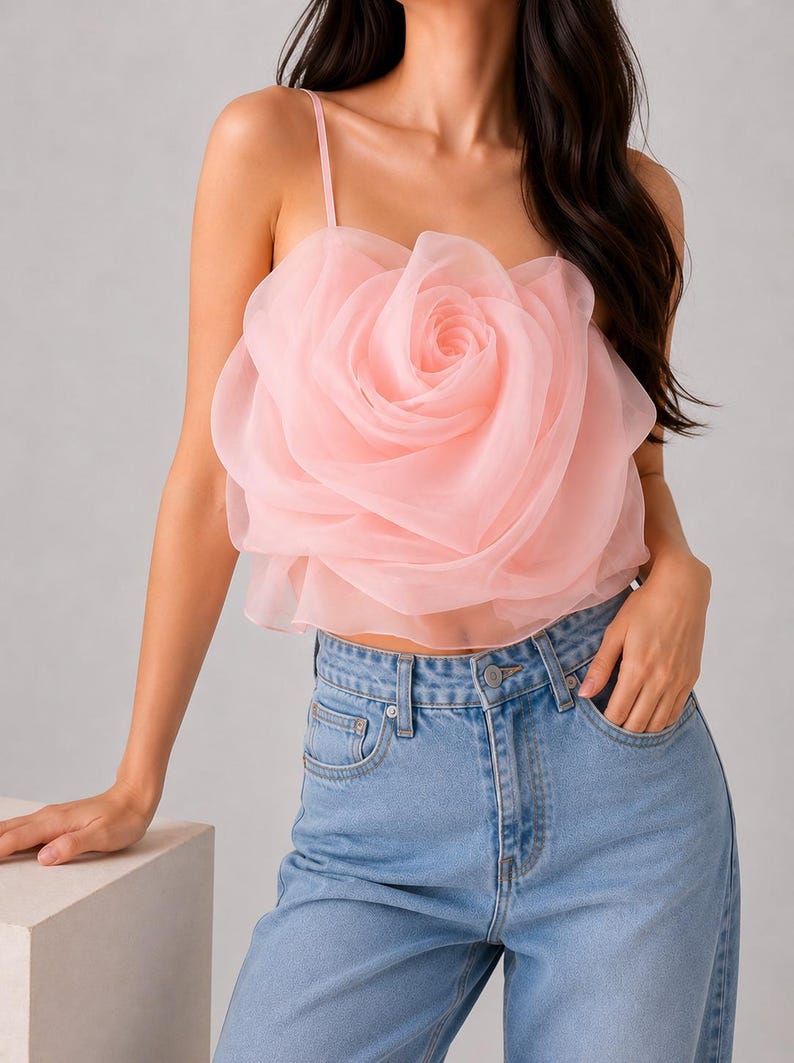 Pu&ograve; includere: Un top corto rosa chiaro con un grande design a forma di rosa realizzato in tessuto a strati. Il top ha sottili spalline ed &egrave; abbinato a jeans di denim blu chiaro.