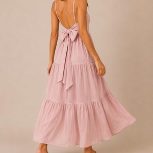 May include: A dusty rose maxi dress featuring a low back design with thin spaghetti straps and a large fabric bow tie at the waist. The garment has a tiered skirt construction with a flowing silhouette that reaches down to the lower calf area.