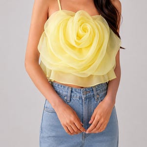 Pu&ograve; includere: Top giallo a forma di fiore con spalline sottili. Il top &egrave; realizzato in un tessuto trasparente e stratificato, creando un effetto rosa tridimensionale. La modella indossa jeans di denim blu chiaro.