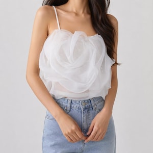 Pu&ograve; includere: Un top bianco senza maniche con un grande design a forma di rosa a strati sul davanti. Il top ha sottili spalline e viene abbinato a jeans di denim blu chiaro. Il tessuto sembra trasparente e delicato.