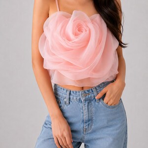 Pu&ograve; includere: Un top corto rosa chiaro con un grande design a rosa a strati. Il top ha sottili spalline e viene abbinato a jeans di denim blu chiaro. Il dettaglio della rosa &egrave; il punto focale del top.