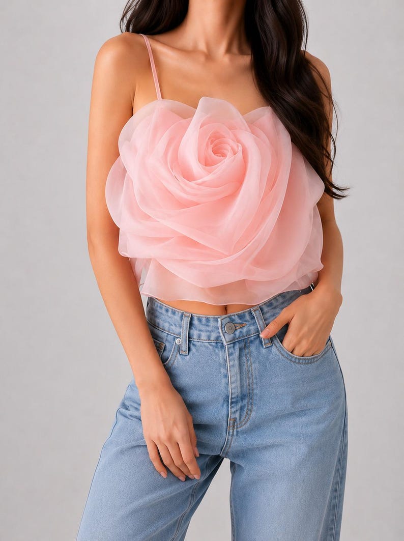 Pu&ograve; includere: Un top corto rosa chiaro con un grande design a rosa a strati. Il top ha sottili spalline e viene abbinato a jeans di denim blu chiaro. Il dettaglio della rosa &egrave; il punto focale del top.