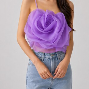 Pu&ograve; includere: Un top a camisole color lavanda con un grande design a rosa tridimensionale. Il top ha sottili spalline e viene abbinato a jeans di denim blu chiaro. La rosa &egrave; il punto focale, creando un look unico e accattivante.