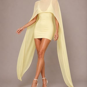May include: A pale yellow mini dress featuring a fitted bodice with a scoop neckline and a sheer, high-neck cape overlay that extends into long, flowing panels on both sides. The garment is paired with simple white strappy high-heeled sandals.
