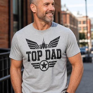 May include: A gray t-shirt with the text "TOP DAD" in black, featuring a stylized jet and star design. The shirt is short-sleeved and made of a soft material.
