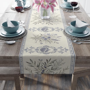 Könnte beinhalten: Ein cremefarbener Tischläufer mit blauen Blumen- und Architekturmustern. Der Läufer liegt auf einem Holztisch, umgeben von Gedecken mit blauen Schalen, Tellern und Weingläsern. Rosa Lilien in einer Glasvase sorgen für einen Farbtupfer.