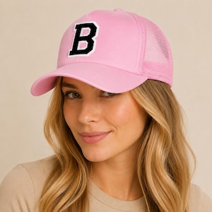 May include: A light pink trucker cap featuring a structured front panel with a large, bold black letter B outlined in white. The cap includes a matching pink mesh back panel and a curved brim, providing a classic athletic look with a vibrant colour.