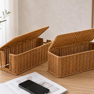May include: Two rectangular woven storage boxes with hinged lids, crafted from light brown synthetic material. Each box features an open side slot for cable management, with a white charging cord visible passing through the opening of one box.