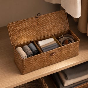 May include: A rectangular brown woven storage box with a hinged lid and three internal compartments. The box contains rolled socks in shades of beige, gray, and charcoal, folded white and gray fabric, and a coiled brown belt with a metal buckle.