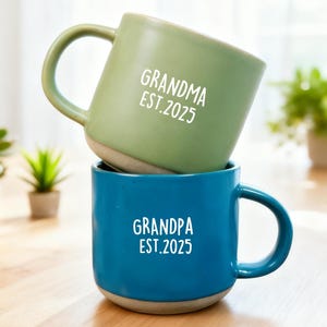 Pode incluir: Duas canecas de cerâmica empilhadas verticalmente. A caneca superior é verde sálvia com o texto branco GRANDMA EST. 2025. A caneca inferior é azul profundo com o texto branco GRANDPA EST. 2025. Ambas têm acabamento liso e base de argila.