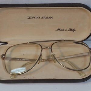 May include: Gold-tone metal aviator eyeglasses with a double bridge design, resting inside a dark brown hard-shell case. The interior of the case features the text Giorgio Armani and Made in Italy printed in black on a light beige lining.