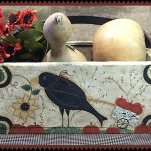 May include: A white painted wooden box with a black crow, sunflowers, and red apples painted on the front. The box has a handle and is filled with gourds.