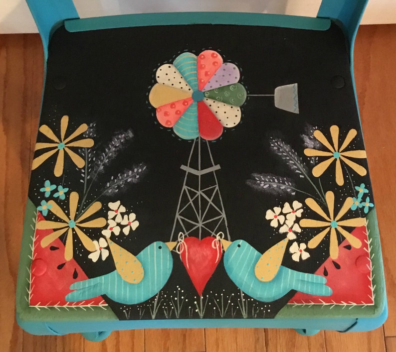 May include: A hand-painted wooden chair with a black seat featuring a whimsical scene of a windmill, two birds, and flowers. The chair is painted in shades of teal, green, and red.