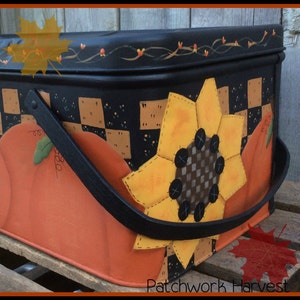 Pu&ograve; includere: Una scatola di metallo nero con un design a patchwork che presenta un girasole e zucche. La scatola ha una maniglia nera e le parole "Patchwork Harvest" sono stampate sul fondo.