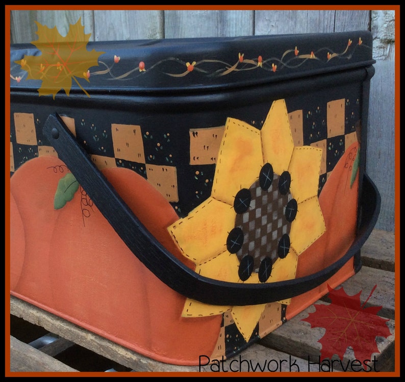 Pu&ograve; includere: Una scatola di metallo nero con un design a patchwork che presenta un girasole e zucche. La scatola ha una maniglia nera e le parole "Patchwork Harvest" sono stampate sul fondo.