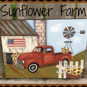 May include: A red vintage pickup truck with a wooden bed filled with sunflowers is parked in front of a white farmhouse with an American flag. A windmill stands in the background with a blue sky and white clouds. The sign on the fence reads "Open".