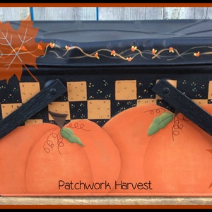 Könnte beinhalten: Schwarzer Metall-Picknickkorb mit kariertem Muster und bemalten Kürbissen. Der Korb hat zwei schwarze Griffe und eine Ranken mit roten Beeren, die obenauf gemalt sind. Der Text "Patchwork Harvest" ist auf der Vorderseite gemalt.