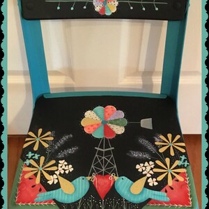Puede incluir: Una silla pintada de turquesa con un asiento negro que presenta un diseño de patchwork caprichoso. El asiento tiene un molino de viento, pájaros y flores. El respaldo de la silla tiene dos paneles negros pintados con un diseño de flores de patchwork. La silla está etiquetada como "Patchwork Garden".