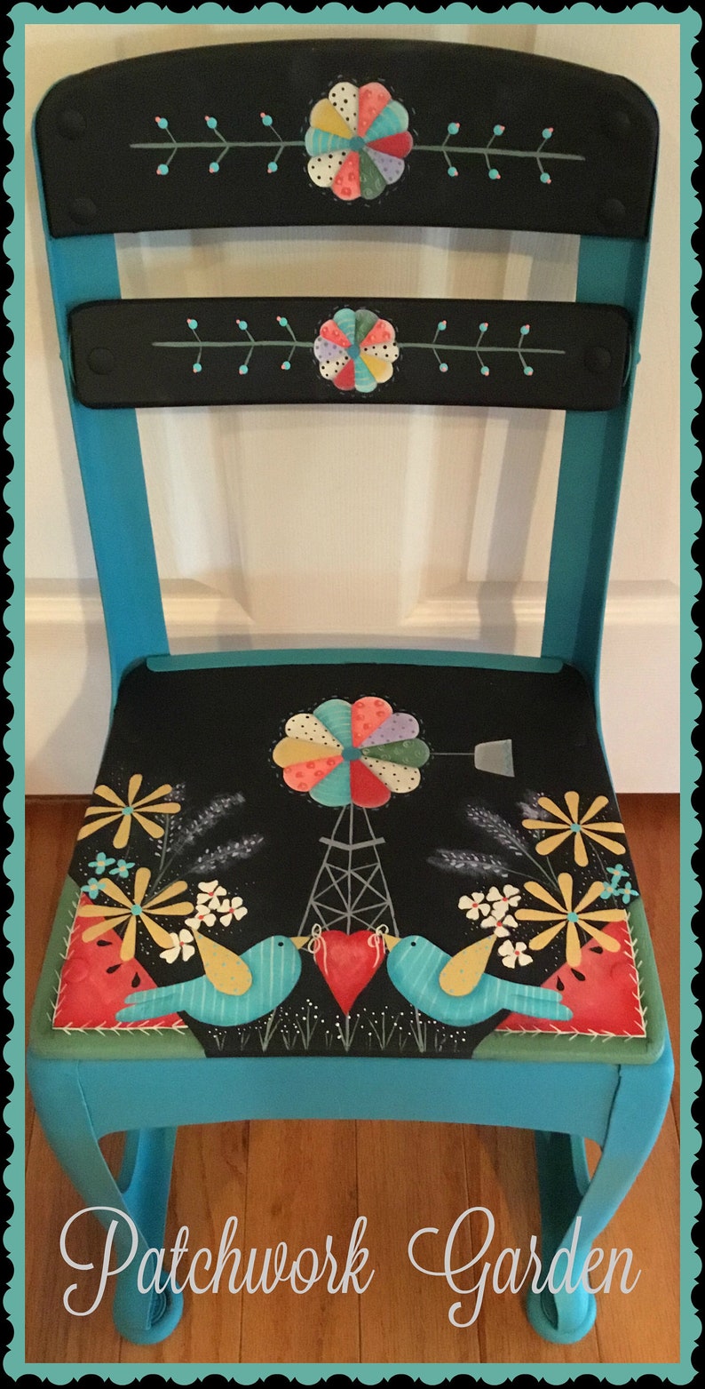 May include: A turquoise painted chair with a black seat featuring a whimsical patchwork design. The seat has a windmill, birds, and flowers. The chair back has two black painted panels with a patchwork flower design. The chair is labelled "Patchwork Garden".