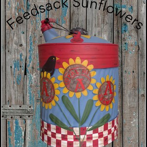 Puede incluir: Un recipiente decorativo de metal pintado con una combinación de colores azul, rojo y amarillo. El recipiente presenta diseños de girasoles y el texto "Feedsack Sunflowers". Un pájaro negro está posado en el lateral, y la base tiene un patrón a cuadros rojo y blanco.