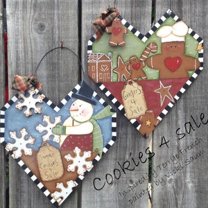 Può includere: Due cartelli di legno a forma di cuore con temi invernali e pan di zenzero dipinti. Il cartello a sinistra presenta un pupazzo di neve che tiene un fiocco di neve e un cartello che dice "Sugar Cookies 4 Sale". Il cartello a destra presenta un uomo di pan di zenzero con un cappello da cuoco e un cartello che dice "Gingers 4 Sale". Il testo "Cookies 4 Sale" è scritto sotto i cartelli. Il testo "Inspired by Terrye French, painted by Vicki Saum" è scritto sotto il testo "Cookies 4 Sale".