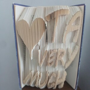 May include: A book sculpture with the words "I LOVE YOU VERY MUCH" carved into the pages. The heart and letters are carefully folded, creating a unique and artistic display. The book has a blue spine and is displayed open.