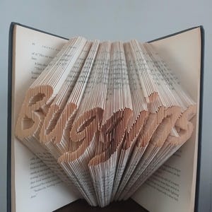 Könnte beinhalten: Ein offenes Buch, dessen Seiten so gefaltet sind, dass das Wort "Believe" entsteht. Das Wort ist aus den Seiten ausgeschnitten, wodurch ein dreidimensionaler Effekt entsteht. Das Buch hat einen dunklen Einband und steht auf einer Holzoberfläche.