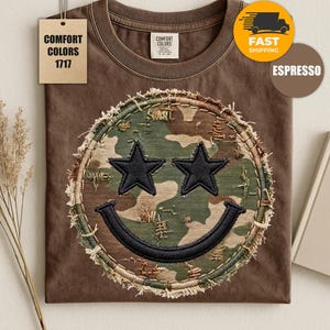 May include: An espresso brown short-sleeve t-shirt featuring a circular patch with a camouflage pattern. The patch has a frayed edge and displays a black embroidered smiley face with two star-shaped eyes and a curved mouth. The shirt has a Comfort Colors 1717 tag.