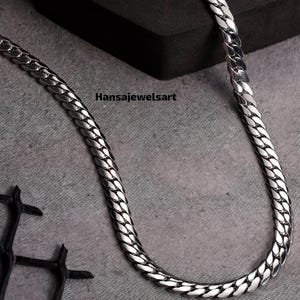 May include: A polished silver-toned curb chain necklace featuring interlocking flat, rounded links with a high-shine finish. The text Hansajewelsart is printed in black near the center of the chain, which is displayed in a curved, U-shaped form.