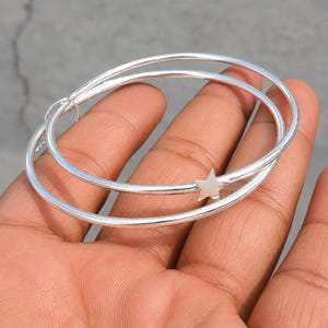 May include: A polished silver-tone bangle bracelet featuring two thin, circular bands that overlap. One of the bands is adorned with a small, five-pointed star charm. The metal has a smooth, reflective finish, creating a clean and simple design.