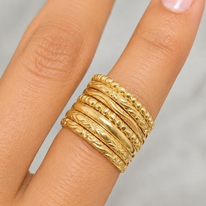 Può includere: Una pila di sei anelli color oro indossati su un dito. Il set include varie texture, con fasce lisce, design a perline e un anello con un delicato motivo inciso, tutti indossati insieme per creare un aspetto stratificato sul dito.