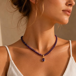 Può includere: Una collana composta da una fila di piccole perle rotonde di ametista viola intenso. La collana presenta un ciondolo centrale color oro con una singola pietra di ametista viola di forma ovale e sfaccettata, incastonata in una montatura liscia.