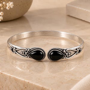 Può includere: Un bracciale aperto in metallo color argento ossidato con due pietre nere a forma di goccia alle estremità. La fascia metallica presenta intricati motivi a volute e dettagli a perline che circondano ogni incastonatura delle pietre nere.