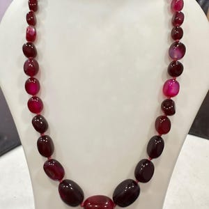 May include: A necklace featuring a single strand of polished, oval-shaped beads in varying shades of deep red, burgundy, and bright magenta. The beads are strung on a thin red cord, with the largest bead positioned at the centre of the strand.