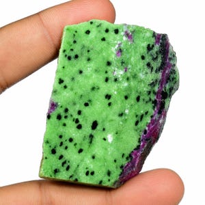 Puede incluir: Una losa plana y en bruto de mineral zoisita que presenta una base de color verde lima vibrante salpicada con numerosas manchas negras pequeñas y una banda distinta e irregular de rubí color magenta profundo en el borde derecho.