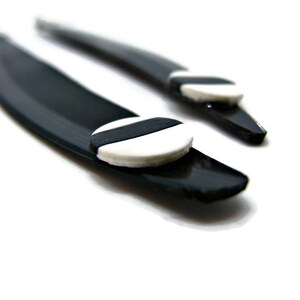 May include: Two black hair barrettes with a curved design. Each barrette features a small, round, white accent with a black stripe. The barrettes are set against a bright white background.