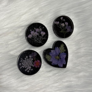 May include: Four decorative resin pieces featuring embedded dried flowers. Three are circular and one is heart-shaped. Each piece has a black background with glitter and contains various pressed purple, pink, and white floral arrangements.
