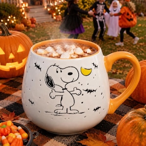 May include: A white ceramic mug with an orange interior and handle, featuring a black line drawing of Snoopy surrounded by small black bats and a yellow crescent moon. The mug is filled with a hot beverage topped with white marshmallows.