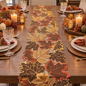 May include: A long table runner featuring an intricate arrangement of embroidered autumn leaves in shades of brown, orange, gold, and yellow. The runner is composed of individual leaf-shaped fabric cutouts layered together to create a textured design.
