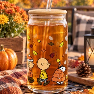 May include: A clear glass tumbler with a bamboo lid and a glass straw, filled with amber liquid. The exterior features a printed graphic of Charlie Brown, Linus, and Snoopy walking amidst falling autumn leaves in shades of yellow, orange, and red.