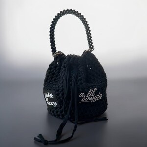 Puede incluir: Un bolso tipo cubo de ganchillo negro con asa de cuentas y cierre de cordón. El bolso presenta dos parches negros con texto bordado en blanco que dice "take it easy" y "a lil' bougie" en las superficies frontal y lateral.