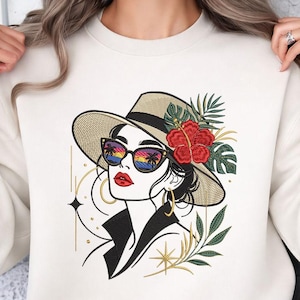 Op de afbeelding: Crèmekleurige sweatshirt met een geborduurde illustratie van een vrouw met een strohoed, zonnebril en rode bloem. De zonnebril weerspiegelt een zonsondergang. Gouden accenten en botanische elementen maken het ontwerp af.