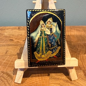 Può includere: Una scatola rettangolare in lacca nera con una scena dipinta a mano di due figure in abiti folcloristici tradizionali. Le figure indossano abiti decorati con dettagli dorati e copricapi. Il bordo è decorato con motivi e fregi dorati.