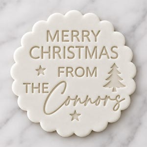 May include: A round white scalloped cookie stamp featuring the embossed text MERRY CHRISTMAS FROM THE Connors, accompanied by a small pine tree icon and two five-pointed star icons, creating a clear and legible holiday message.