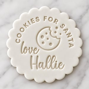 May include: A round white cookie stamp with a scalloped edge featuring the embossed text "COOKIES FOR SANTA" in a sans-serif font, a graphic of a bitten chocolate chip cookie, and the cursive script "love Hallie" centered below.