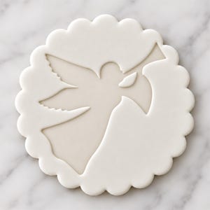 May include: A circular white ceramic coaster with a scalloped edge featuring a recessed, matte-finish silhouette of a dove in flight. The central dove design is smooth and contrasts with the slightly textured white border of the coaster.