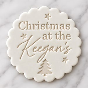 May include: A white circular cookie stamp with a scalloped edge featuring the embossed text "Christmas at the Keegan's" in a mix of serif and script fonts, accented by three small stars and a stylized pine tree at the bottom.