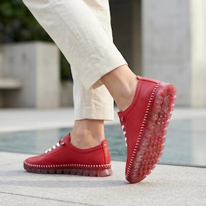 Puede incluir: Zapatos de cuero rojo con cordones y costuras blancas. Las suelas son transparentes, revelando el dibujo de la banda de rodadura. Los zapatos se usan con pantalones color crema.