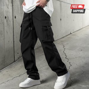 Könnte beinhalten: Schwarze Cargohose mit mehreren Taschen und einem Kordelzug in der Taille. Die Hose wird mit einem weißen T-Shirt und weißen Turnschuhen getragen. Das Bild ist in einer urbanen Umgebung aufgenommen.